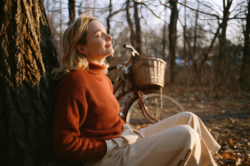 Young blonde woman resting against a tree in a forest with a vintage bicycle nearby during autumn