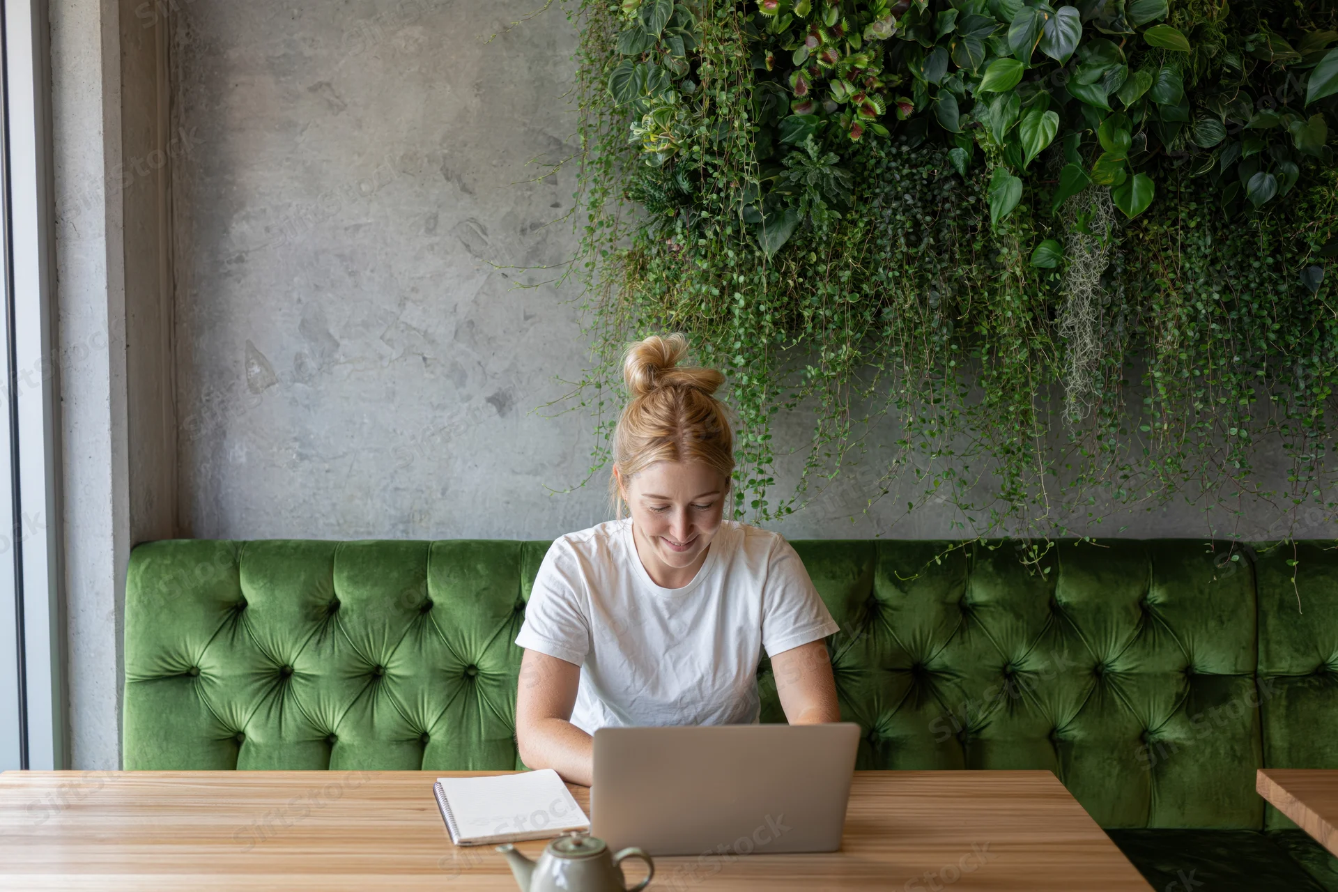 Young blonde woman working on laptop at wooden cafe table with green plants behind her