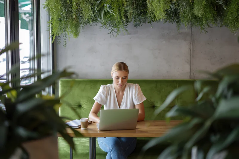 Woman working on laptop at wooden cafe table surrounded by green plants