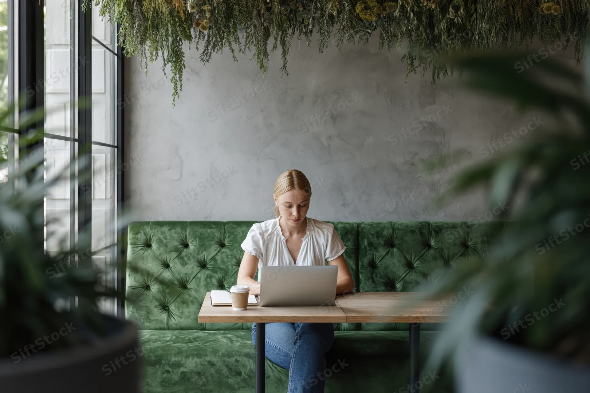 Young blonde woman working on laptop at wooden cafe table with green sofa and plants around