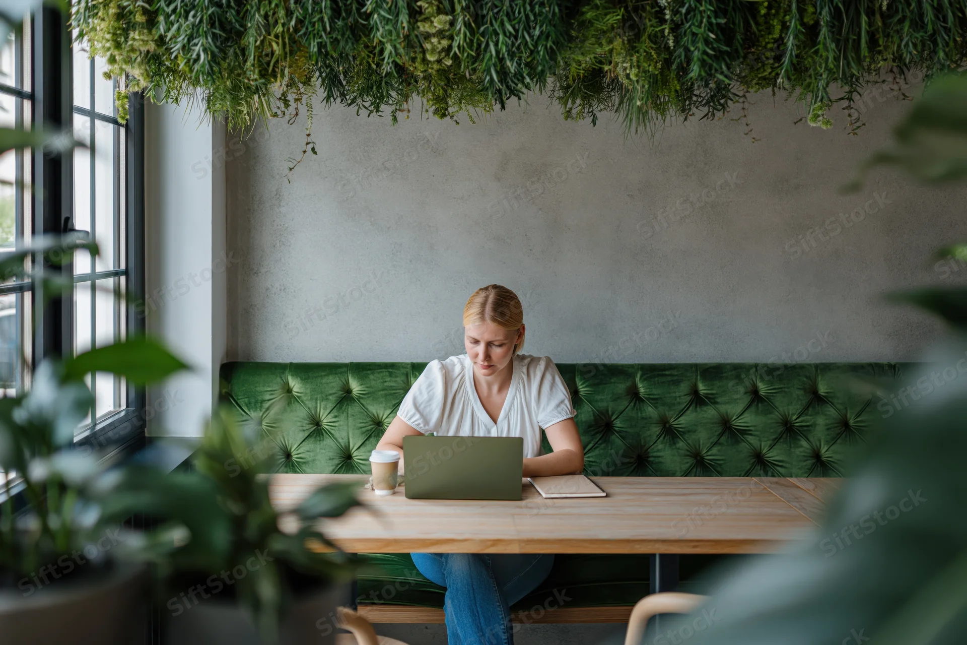 Young blonde woman working on laptop at wooden cafe table with green plants overhead