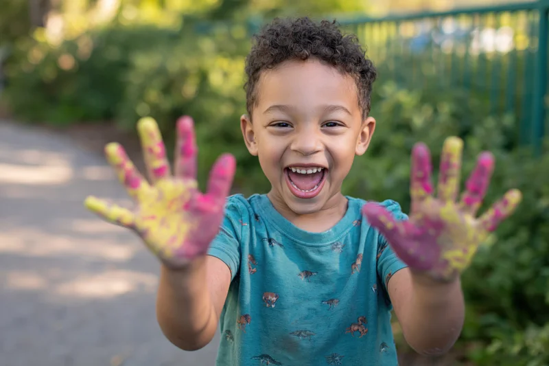 Young boy laughing with paint-covered hands outdoors in a garden
