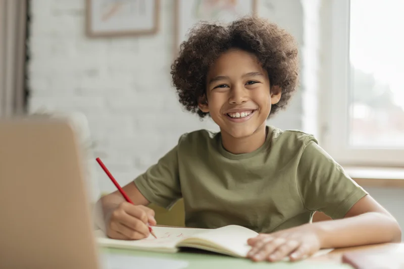 Young boy smiling while writing in a notebook at a desk with a laptop nearby