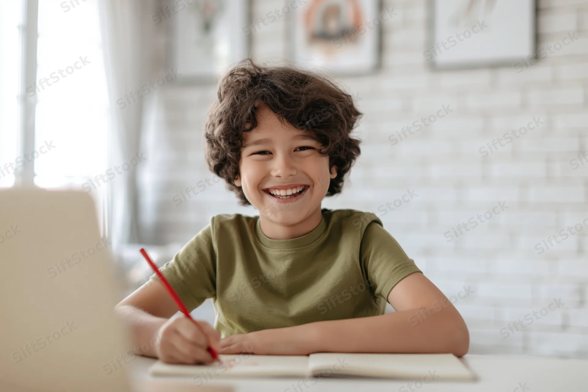 Young boy with curly dark hair laughing while writing in a notebook at a desk at home