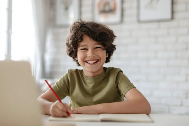 Young boy with curly dark hair laughing while writing in a notebook at a desk at home