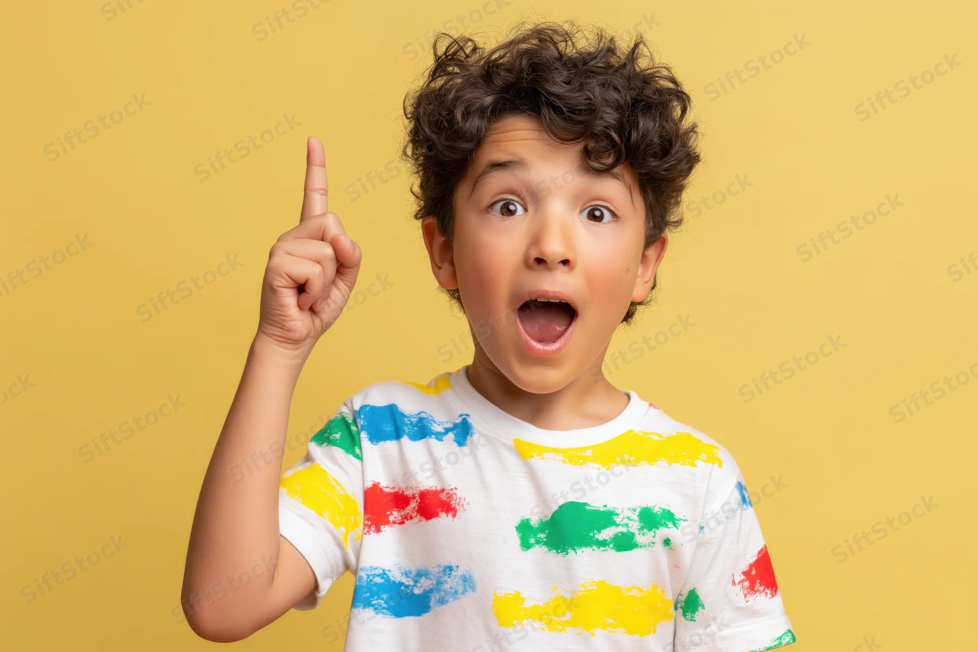 Young boy with curly dark hair pointing upward with surprised expression in colorful striped tshirt against yellow background