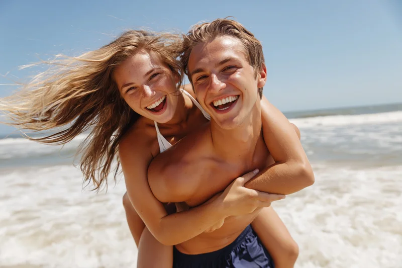 Young couple laughing while piggybacking on a sandy beach near the ocean