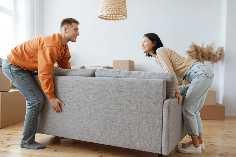 Young couple lifting a gray fabric sofa together in their new apartment