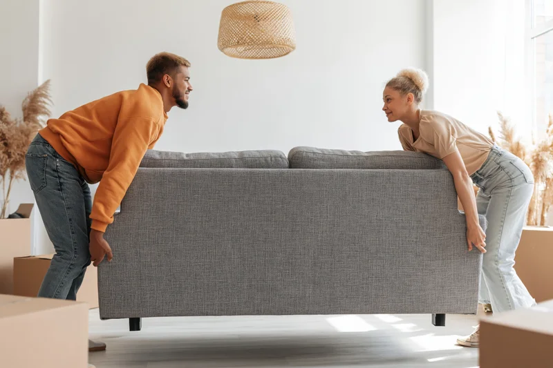 Young couple lifting a gray fabric sofa together in their new apartment