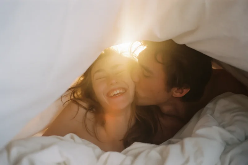 Young couple laughing and kissing under white duvet in bed with sunlight streaming through