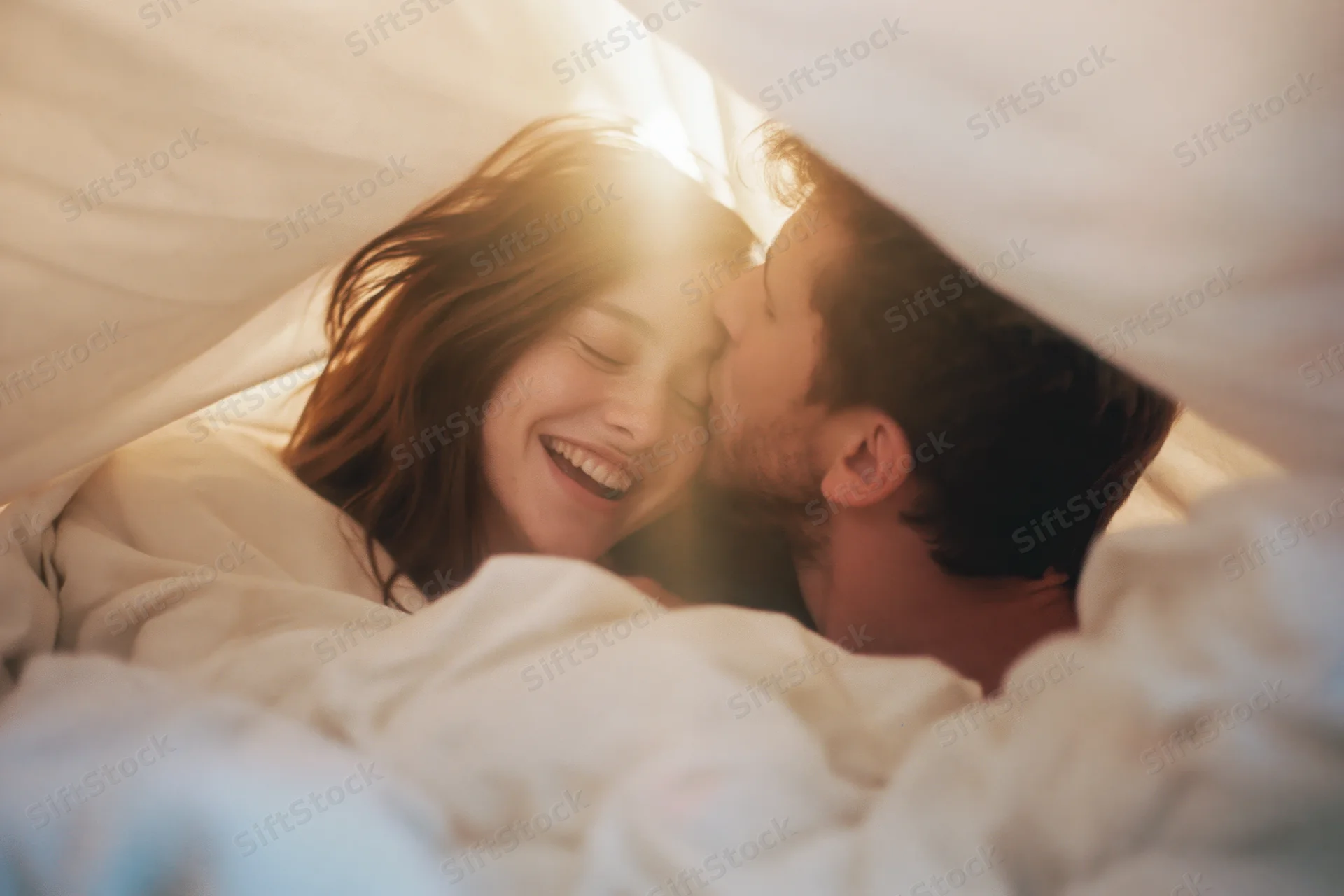 Young couple kissing and smiling under white duvet in bed