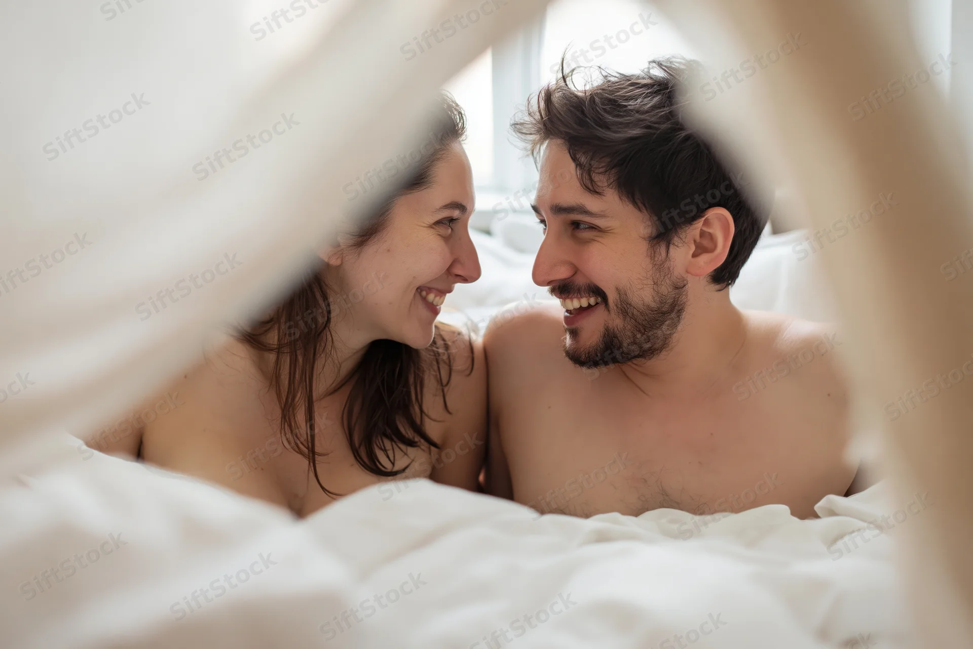Young couple smiling at each other under white sheets in bed