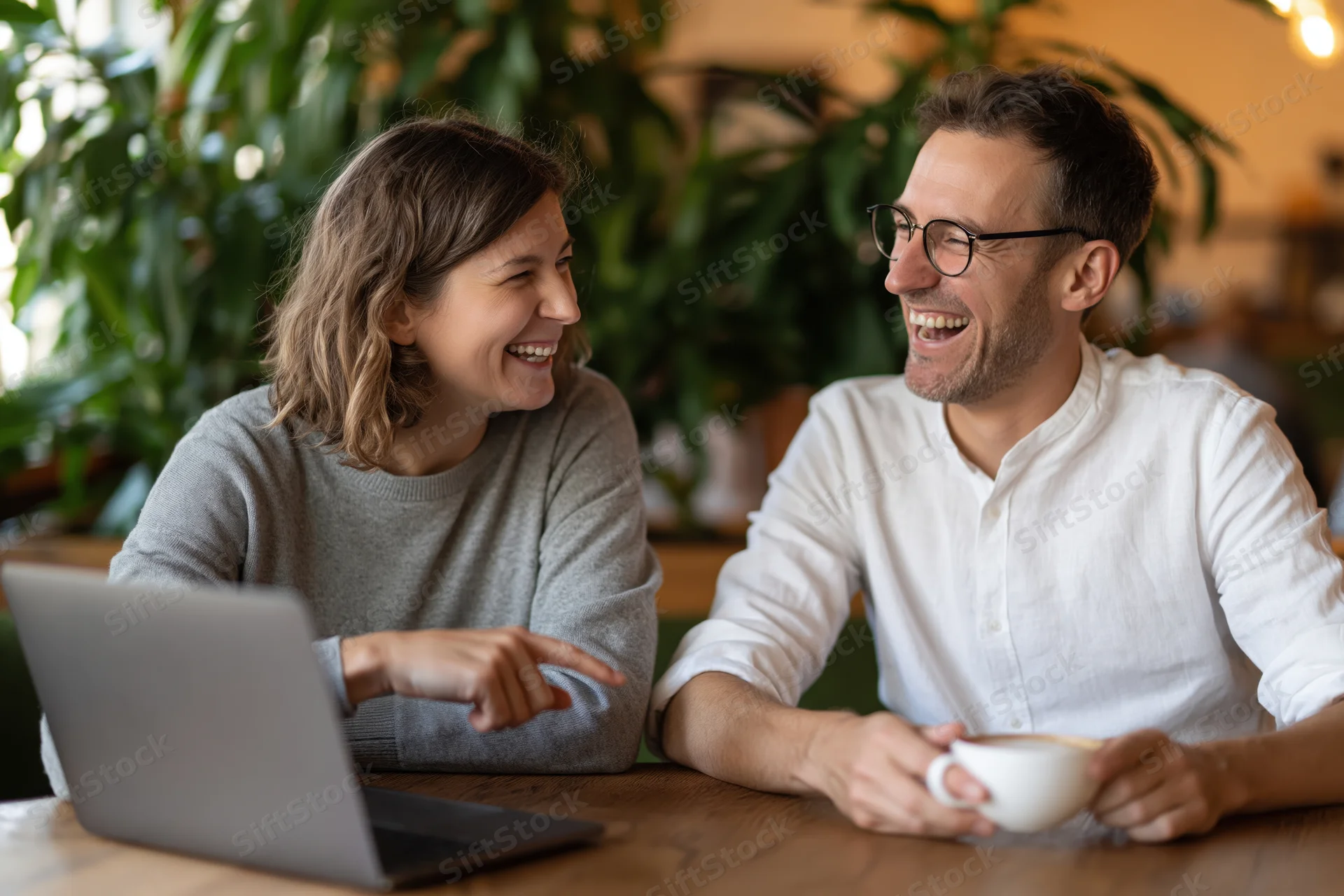 Young couple laughing and talking at a wooden cafe table with laptop and coffee cup