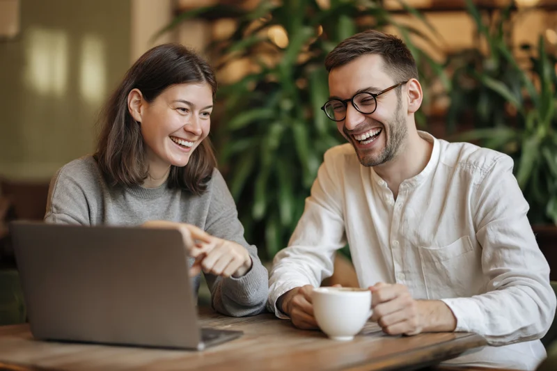 Young couple laughing together at a wooden cafe table with laptop and coffee