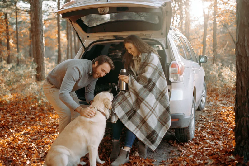 Couple with dog sitting in open trunk of white SUV in autumn forest