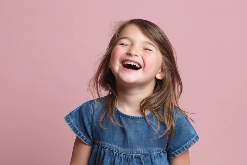 Young girl laughing joyfully in a denim dress against a pink background