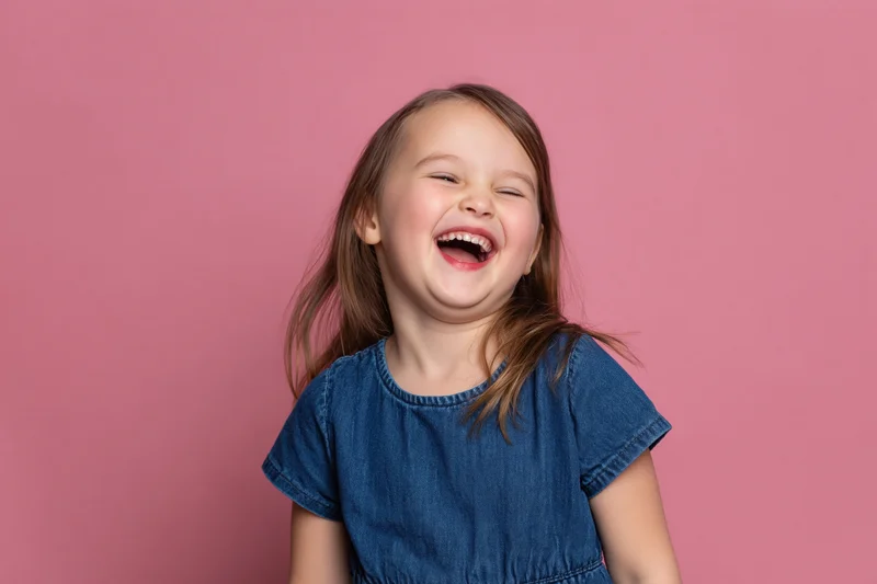 Young girl laughing joyfully in a denim dress against a pink background