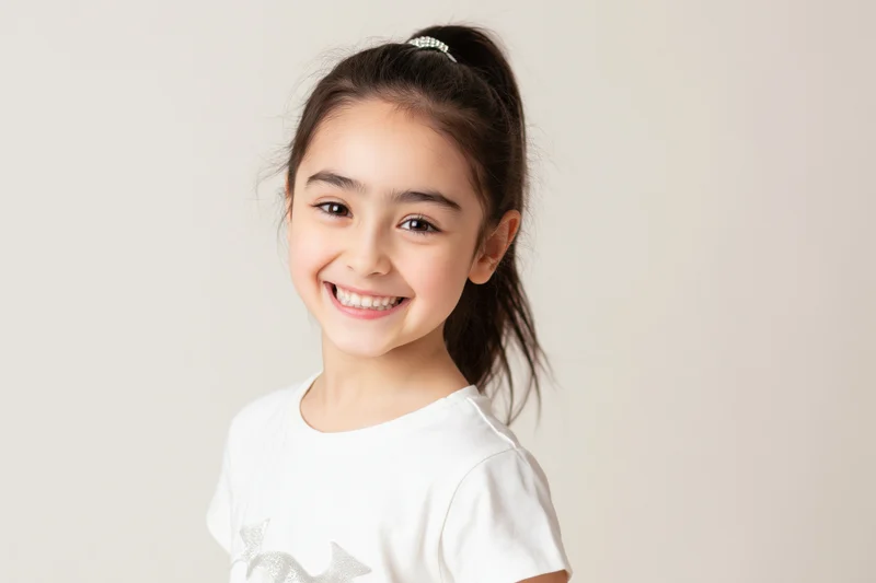 Young girl smiling widely at the camera in a white tshirt with a ponytail against a plain background