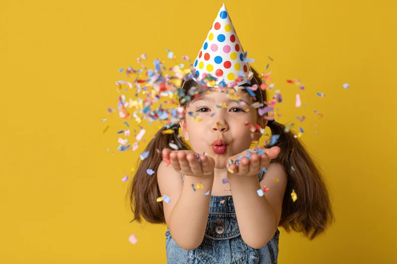 Young girl blowing confetti from her hands while wearing a polka-dot party hat against a yellow background