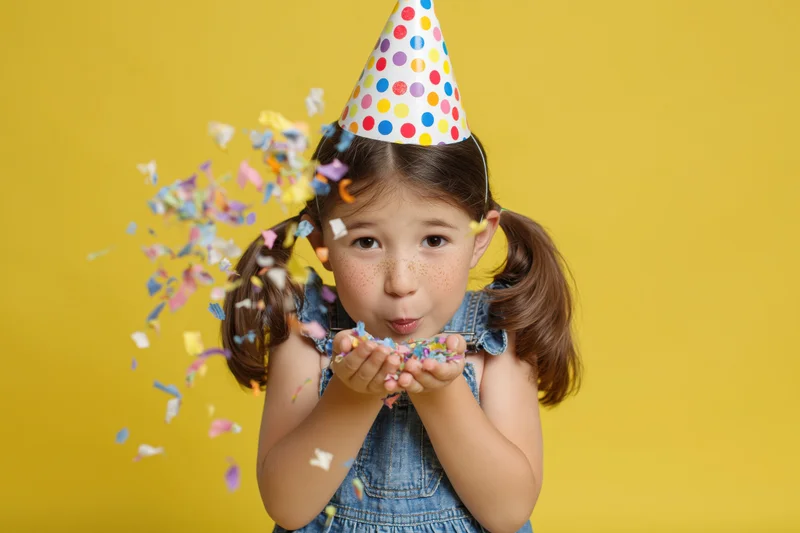 Young girl blowing confetti from her hands while wearing a polka-dot party hat against a yellow background