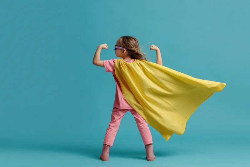 Young girl in superhero costume flexing arms with yellow cape flowing against blue background