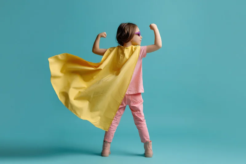 Young girl in superhero costume flexing arms with yellow cape flowing against blue background