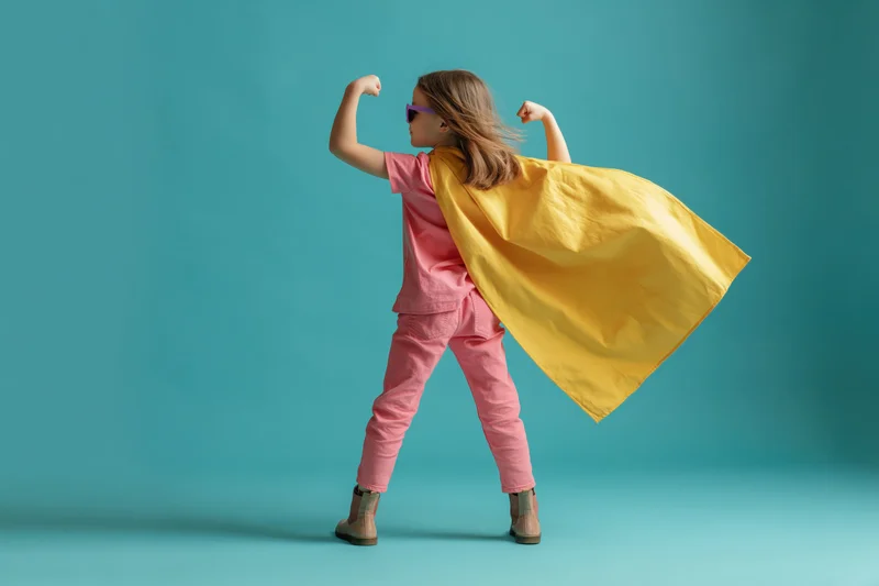Young girl in superhero costume flexing arms with yellow cape flowing in wind against blue background