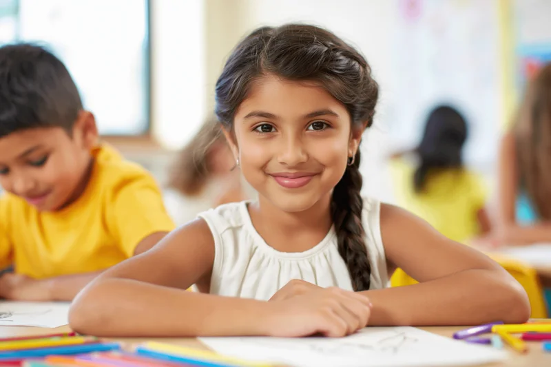 Young girl smiling at camera while drawing at her desk in a classroom