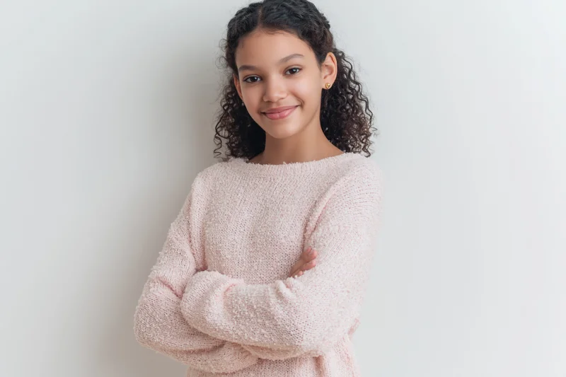 Young girl with curly dark hair smiling at the camera arms crossed in pink knit sweater against white background