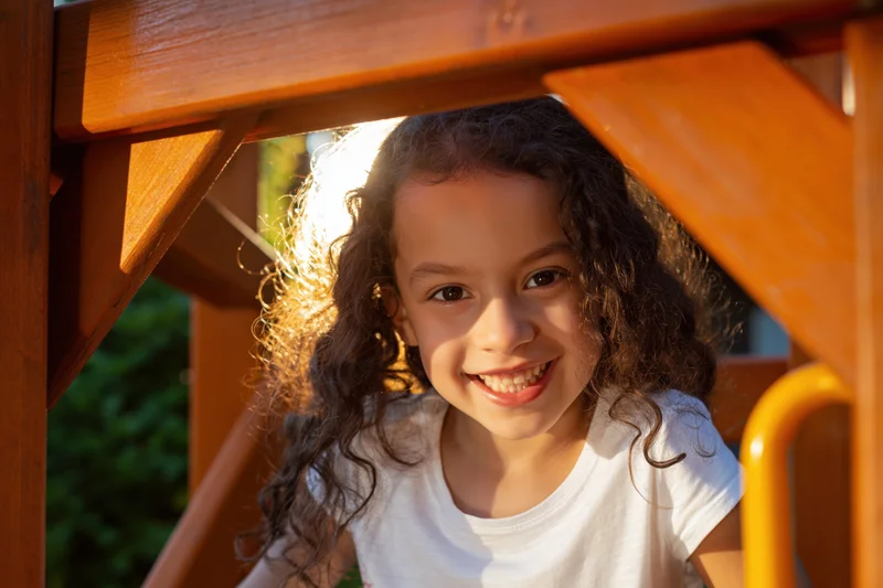 Young girl with curly dark hair smiling widely while peeking through wooden playground structure