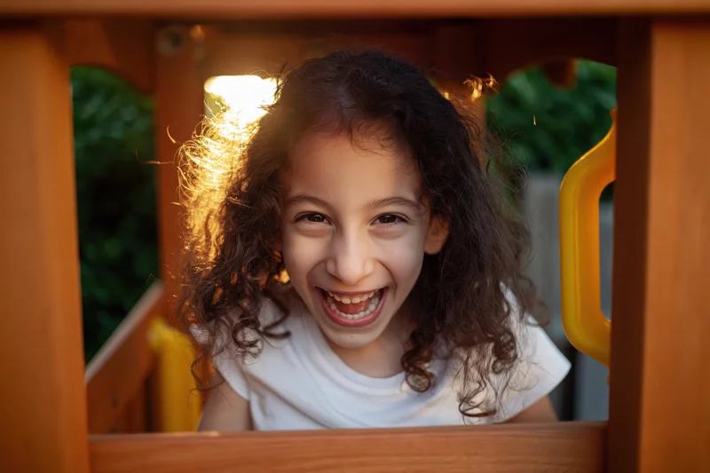 Girl smiling widely while peeking through wooden playground structure