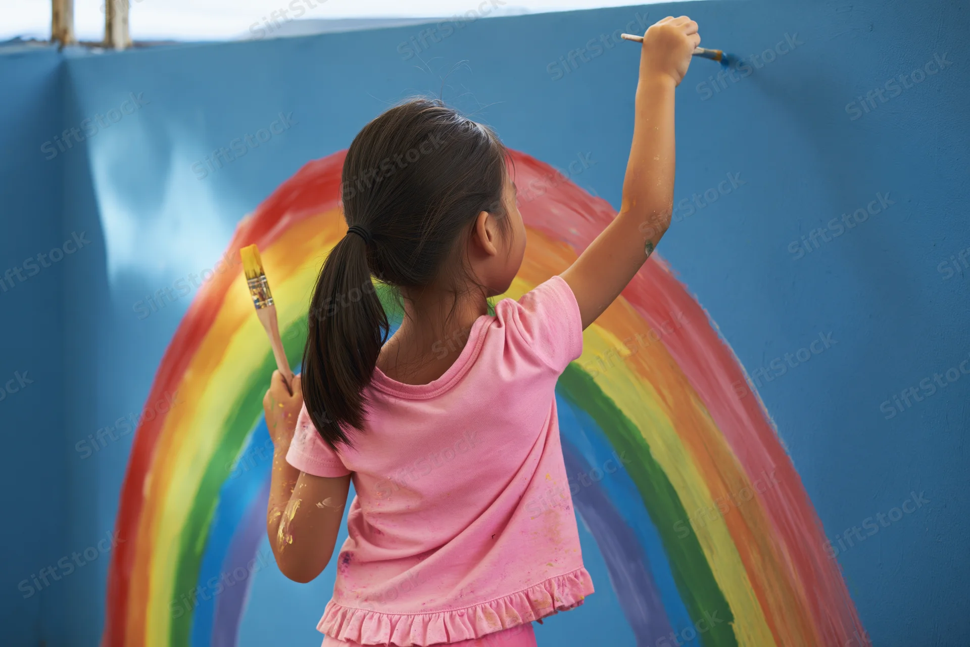 Young girl painting a rainbow on a blue wall outdoors
