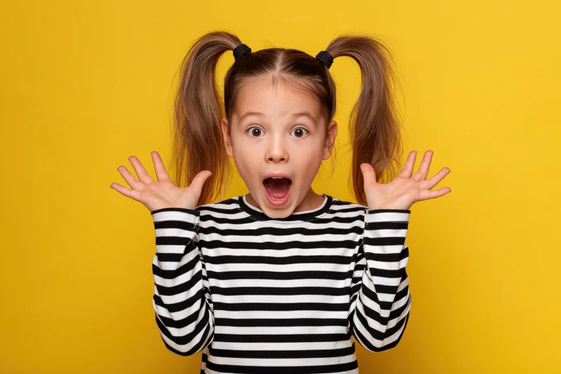 Young girl with high ponytails making surprised expression with hands raised against yellow background