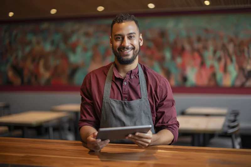Smiling man in apron holding tablet at wooden bar counter in restaurant interior