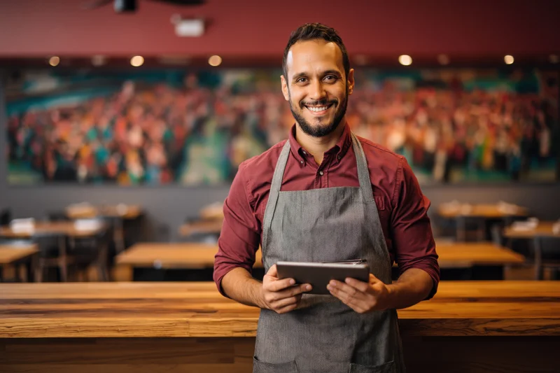 Smiling male restaurant owner holding tablet beside wooden bar counter in dining interior