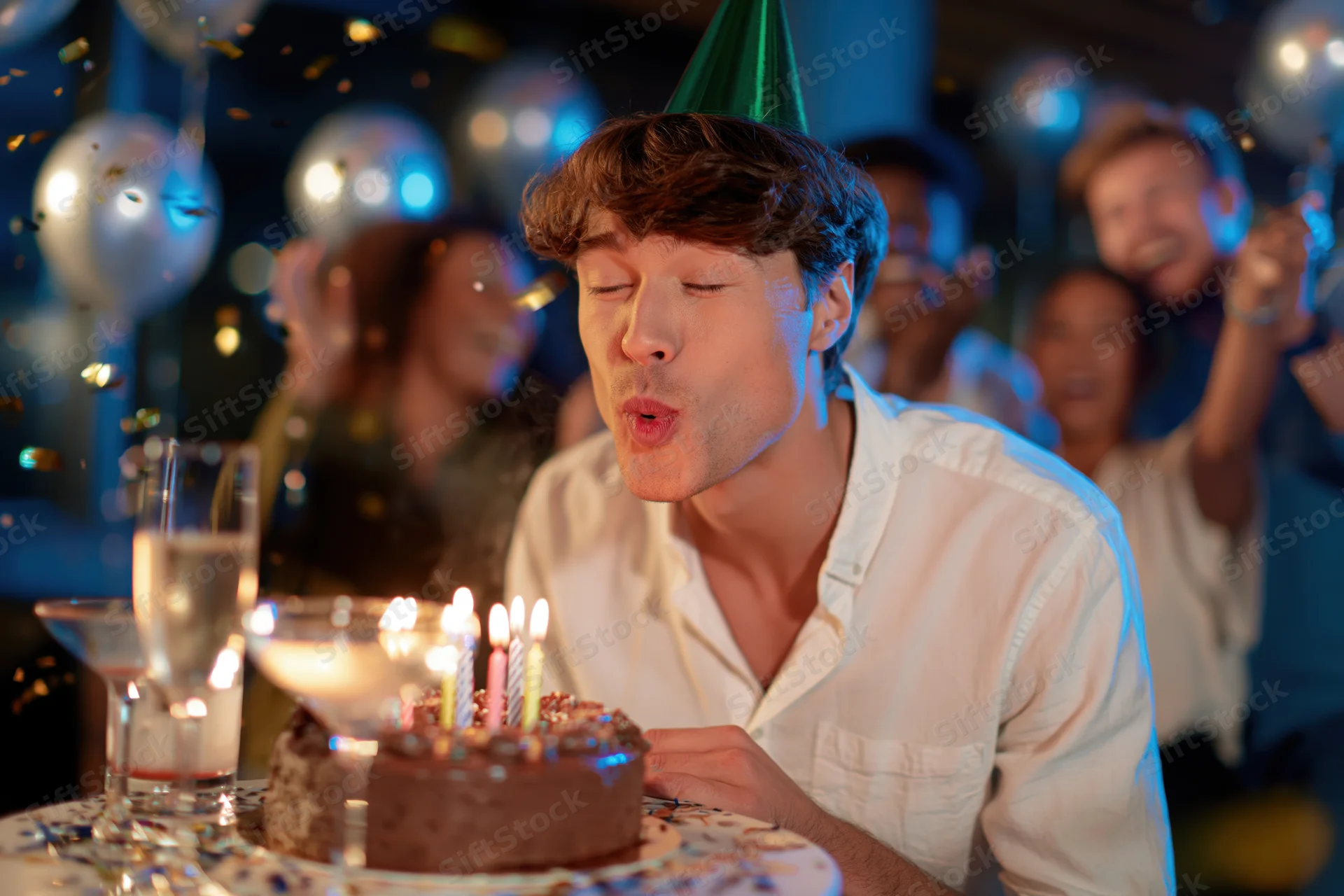 Young man blowing out candles on a chocolate birthday cake at a party with friends and decorations