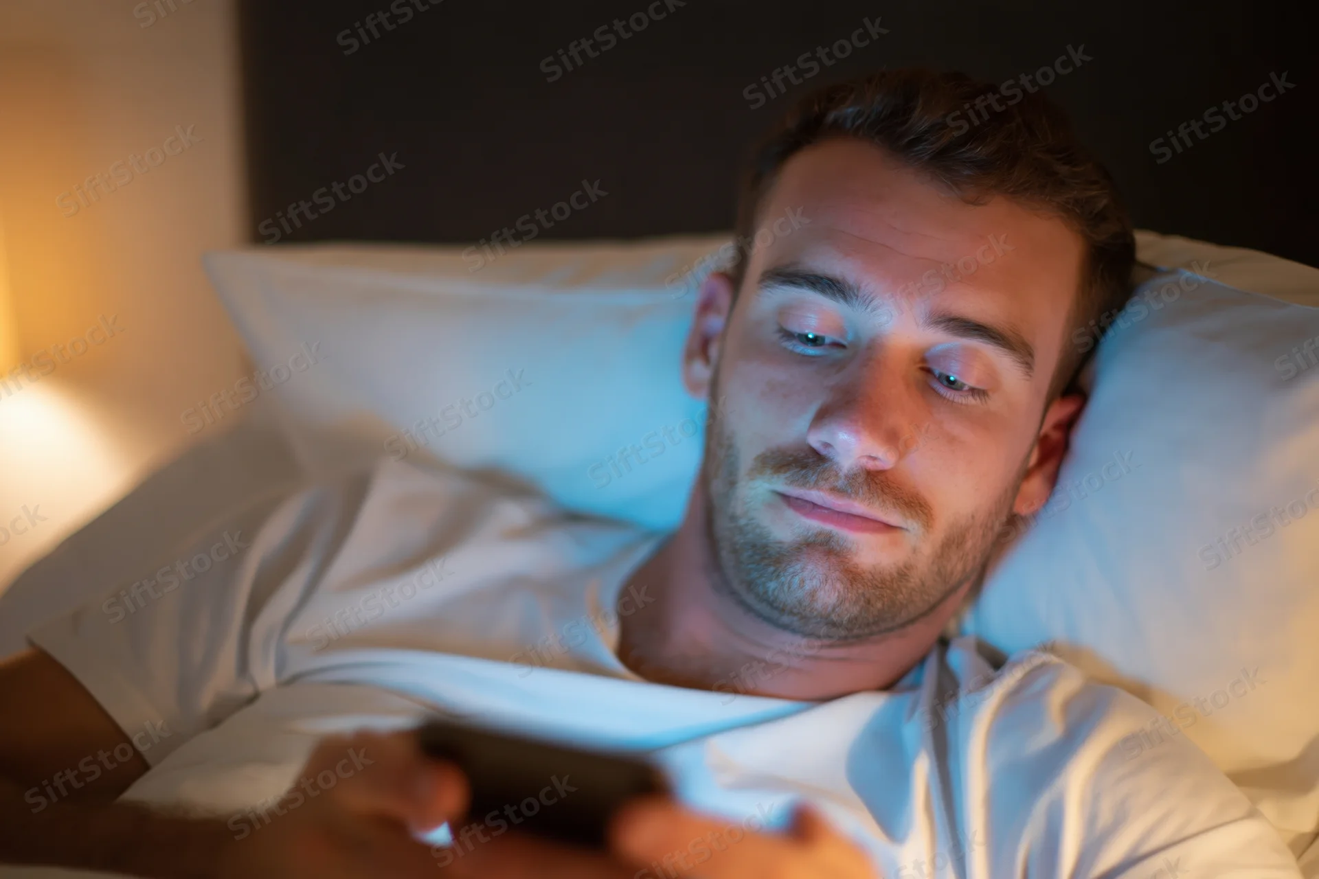 Young man lying in bed at night using smartphone with head on white pillow