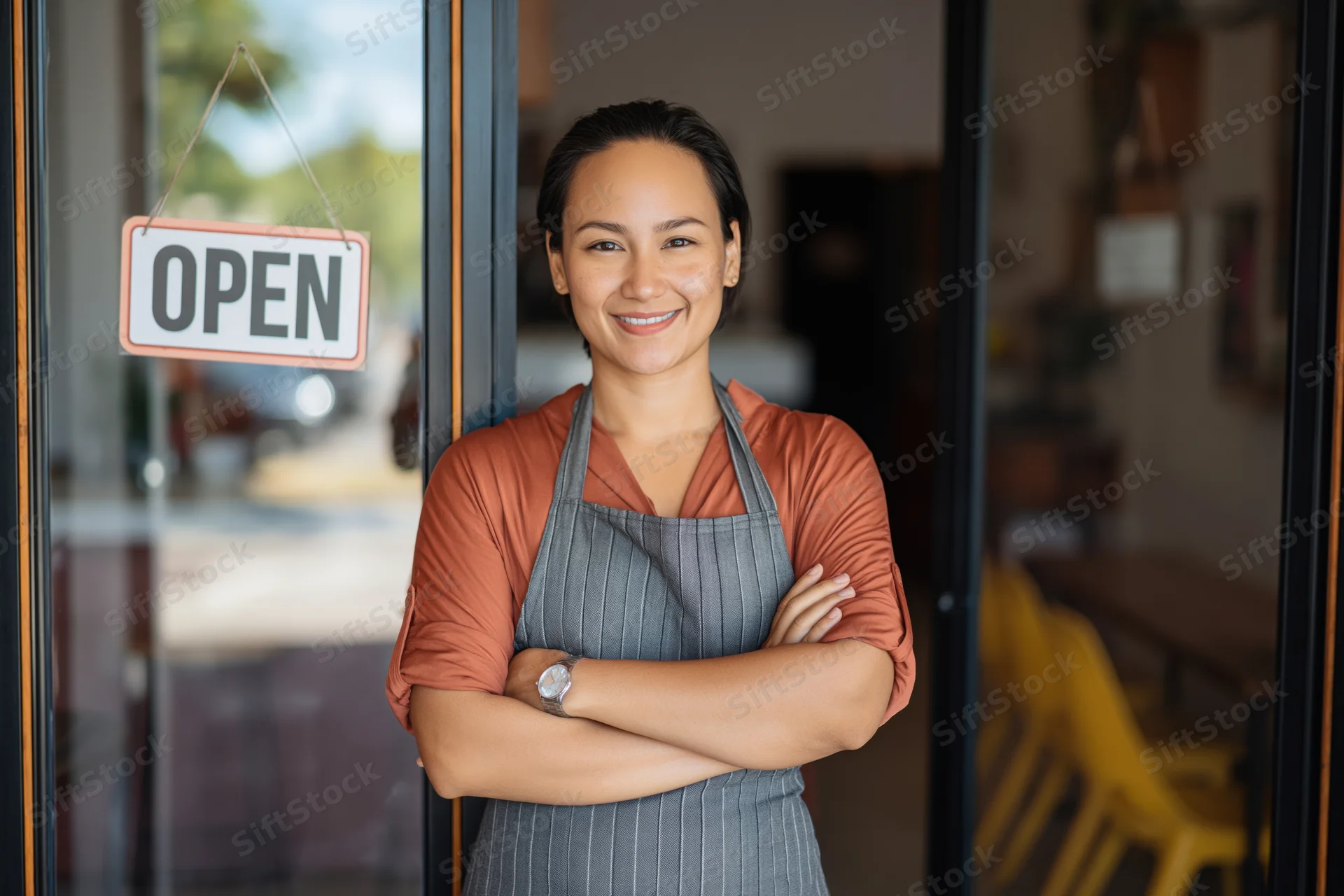 Woman in apron standing at cafe entrance with arms crossed smiling beside open sign