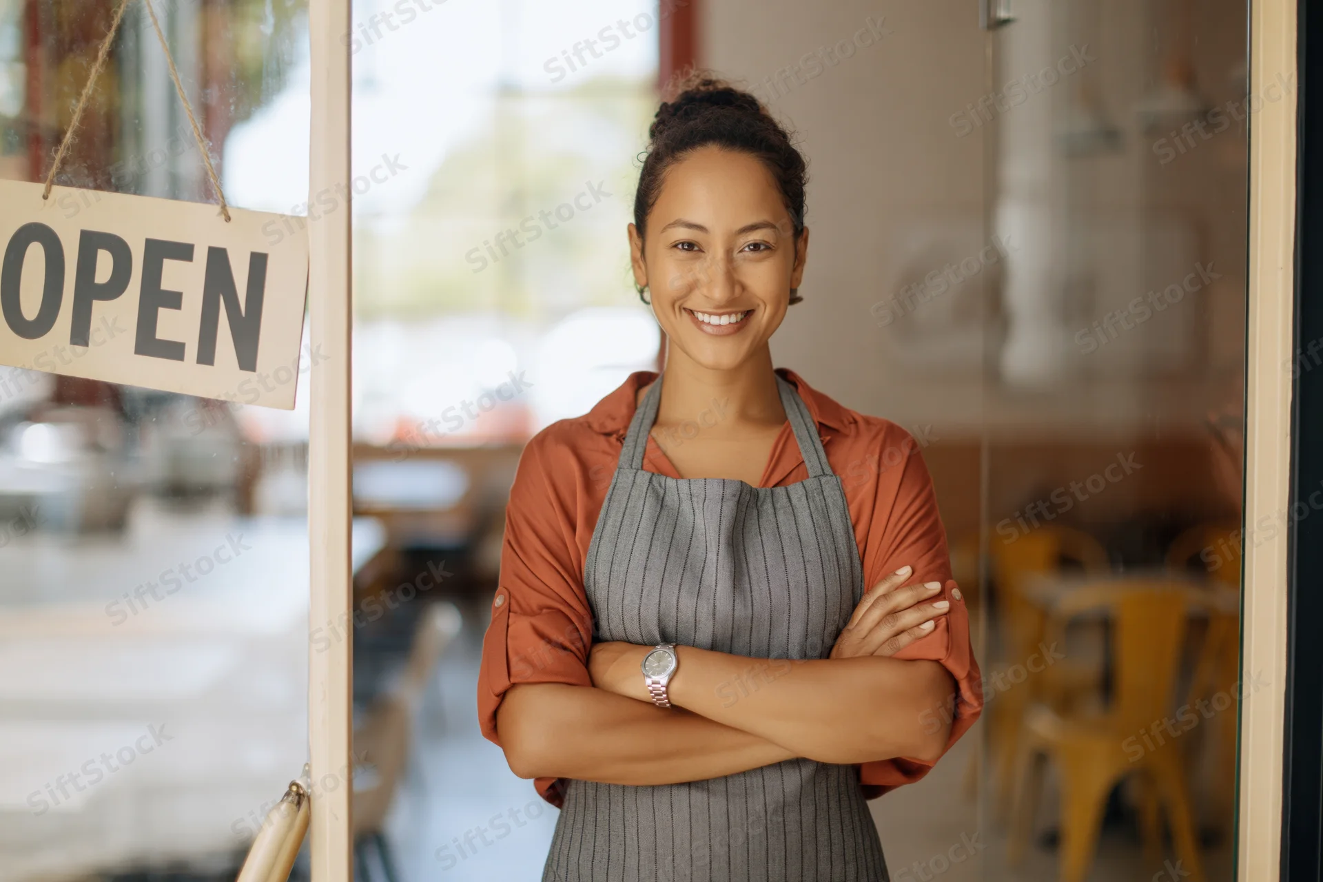 Woman in apron standing at cafe entrance with arms crossed, smiling beside open sign