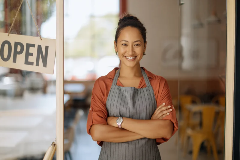 Woman in apron standing at cafe entrance with arms crossed, smiling beside open sign