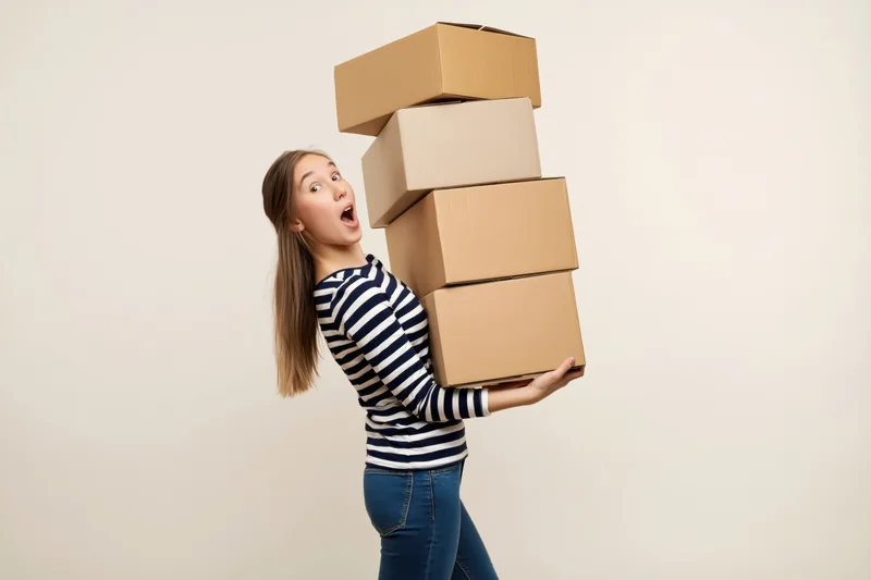 Young woman balancing a tall stack of cardboard boxes while looking surprised