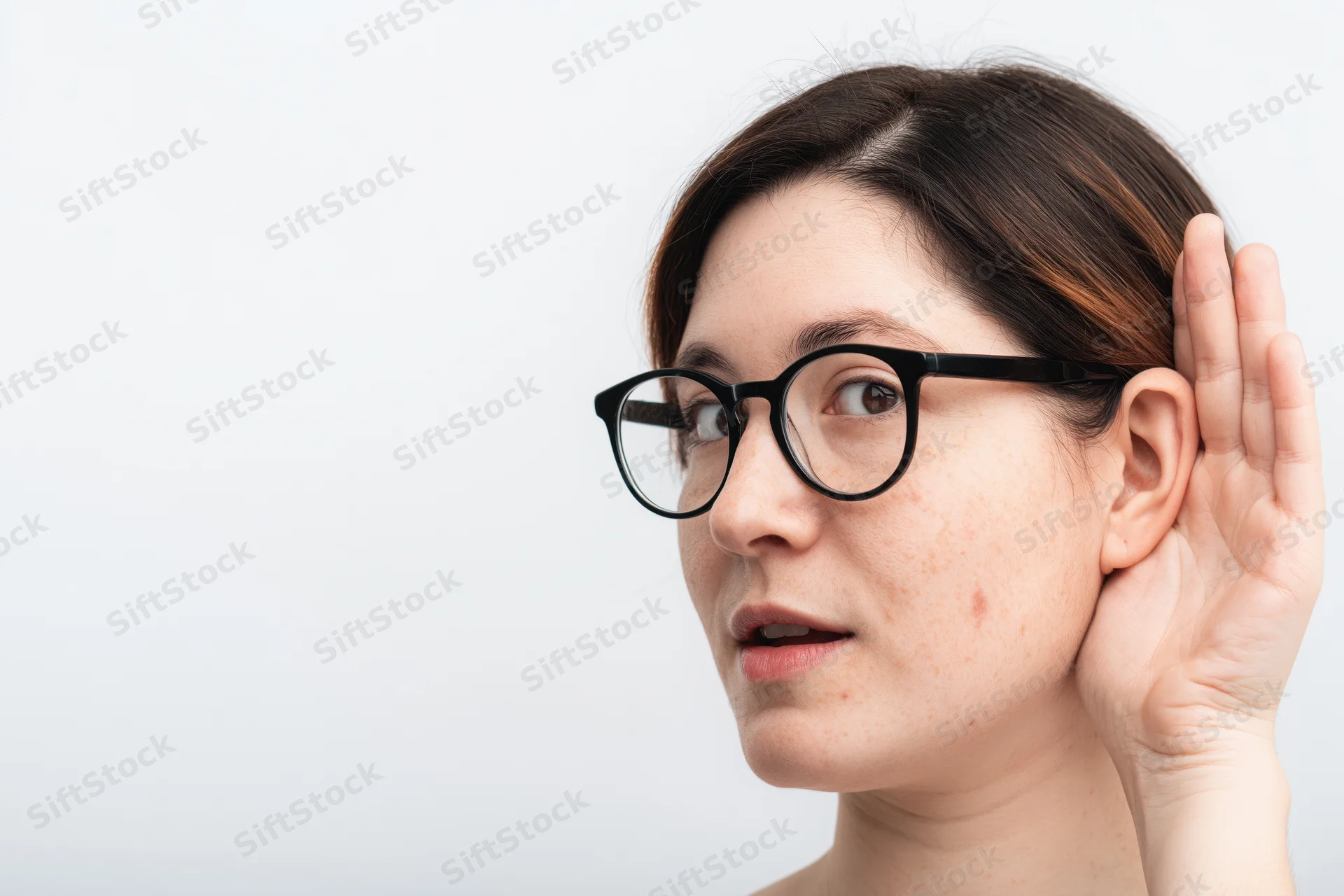 Woman cupping hand behind ear while wearing glasses in studio setting