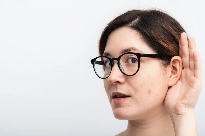 Woman cupping hand behind ear while wearing glasses in studio setting