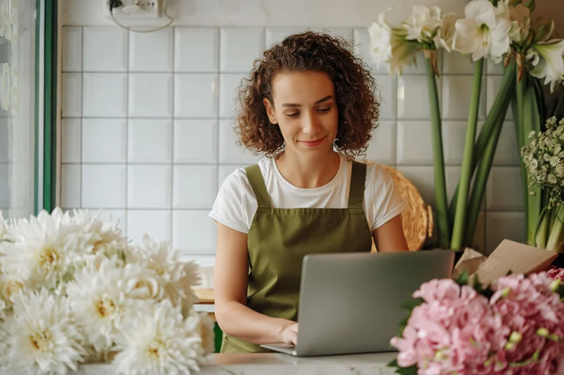 Young woman florist working on laptop behind shop counter surrounded by flowers