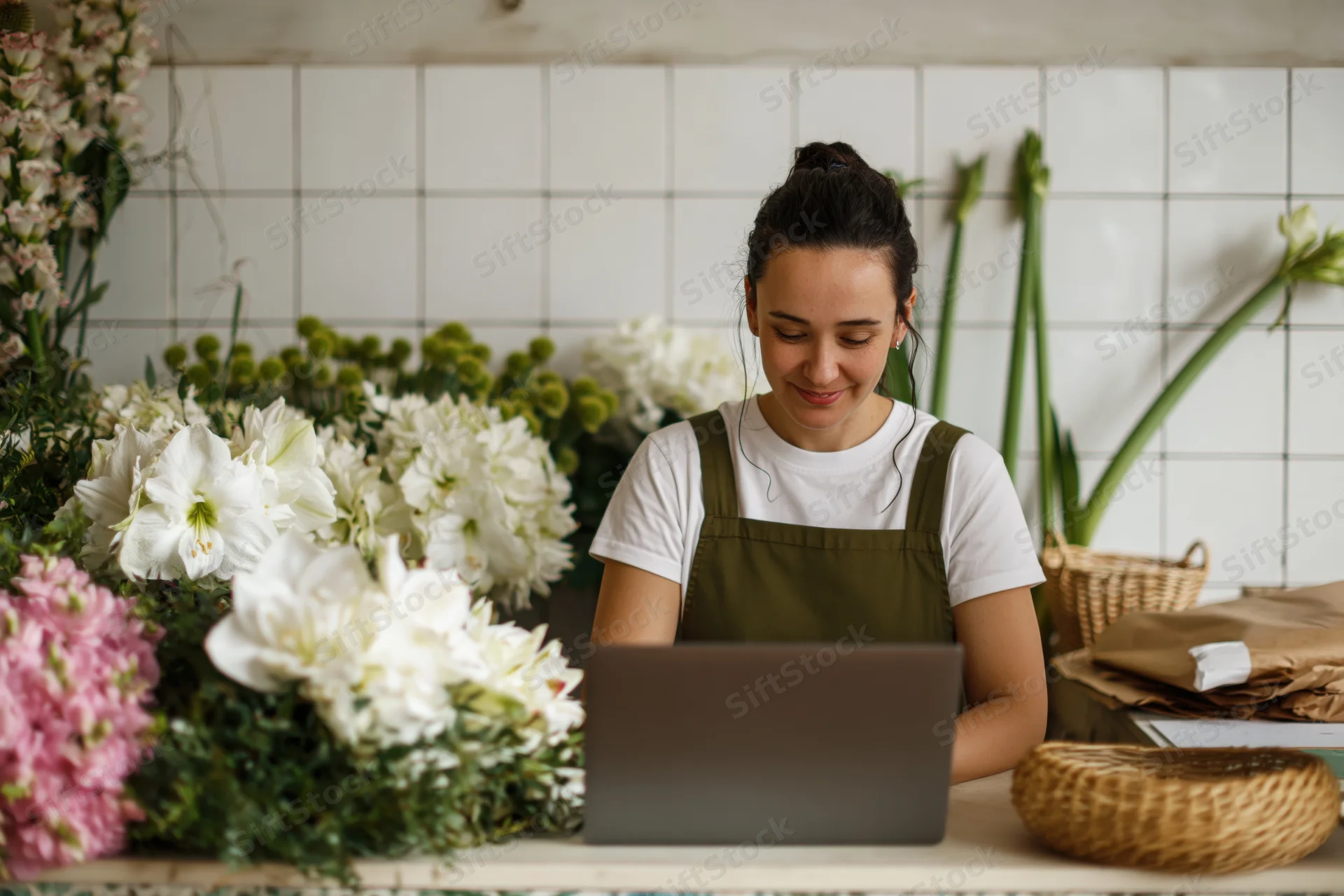 Young woman florist working on laptop behind shop counter surrounded by flowers