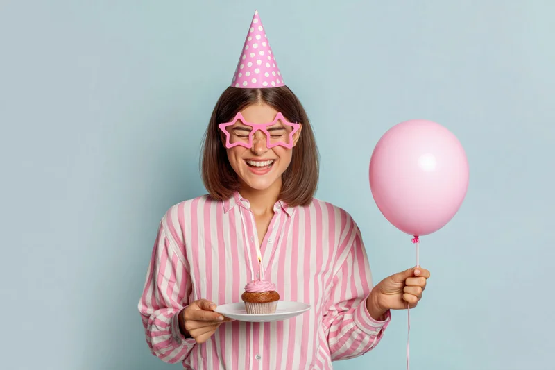 Young woman smiling with eyes closed holding a cupcake and pink balloon, wearing party hat and star sunglasses