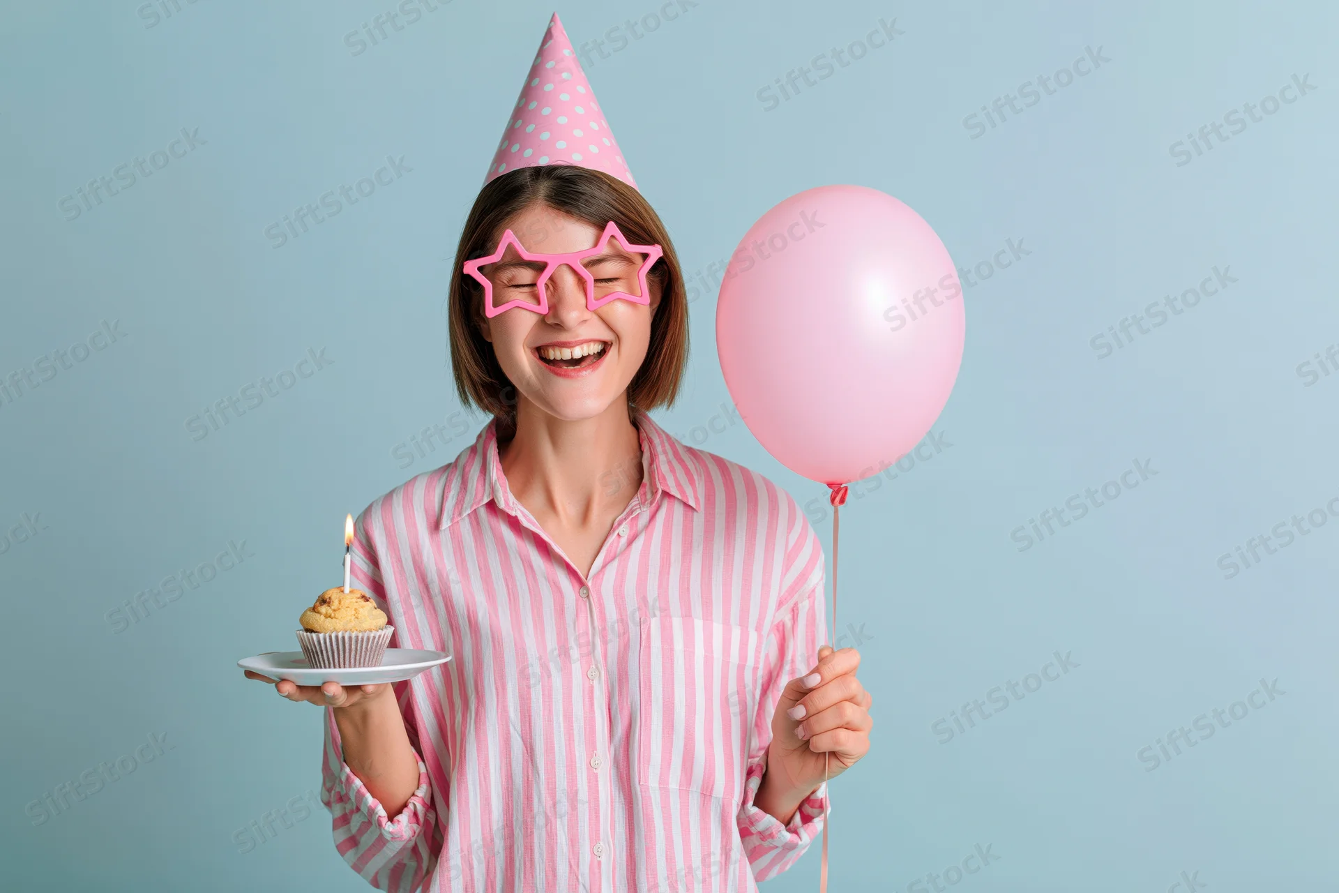 Young woman smiling with eyes closed holding a cupcake and pink balloon, wearing party hat and star glasses against light blue background
