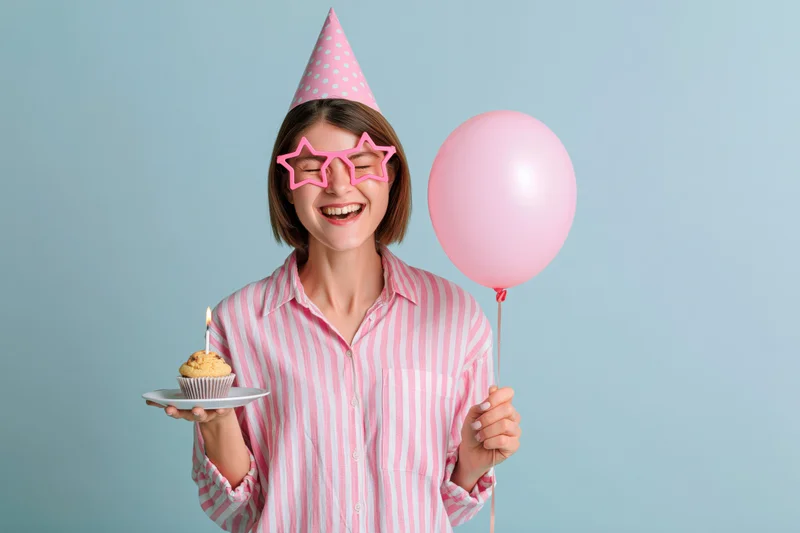 Young woman smiling with eyes closed holding a cupcake and pink balloon, wearing party hat and star glasses against light blue background