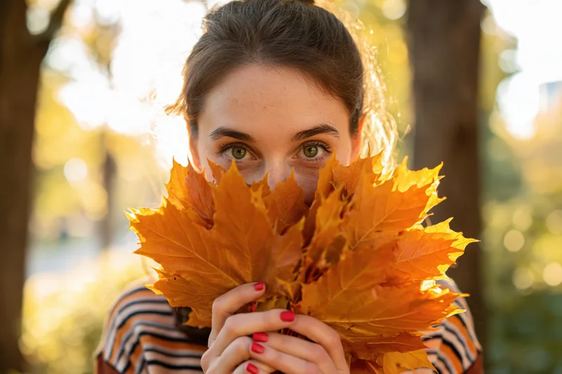Young woman holding orange maple leaves over her face in a sunlit park