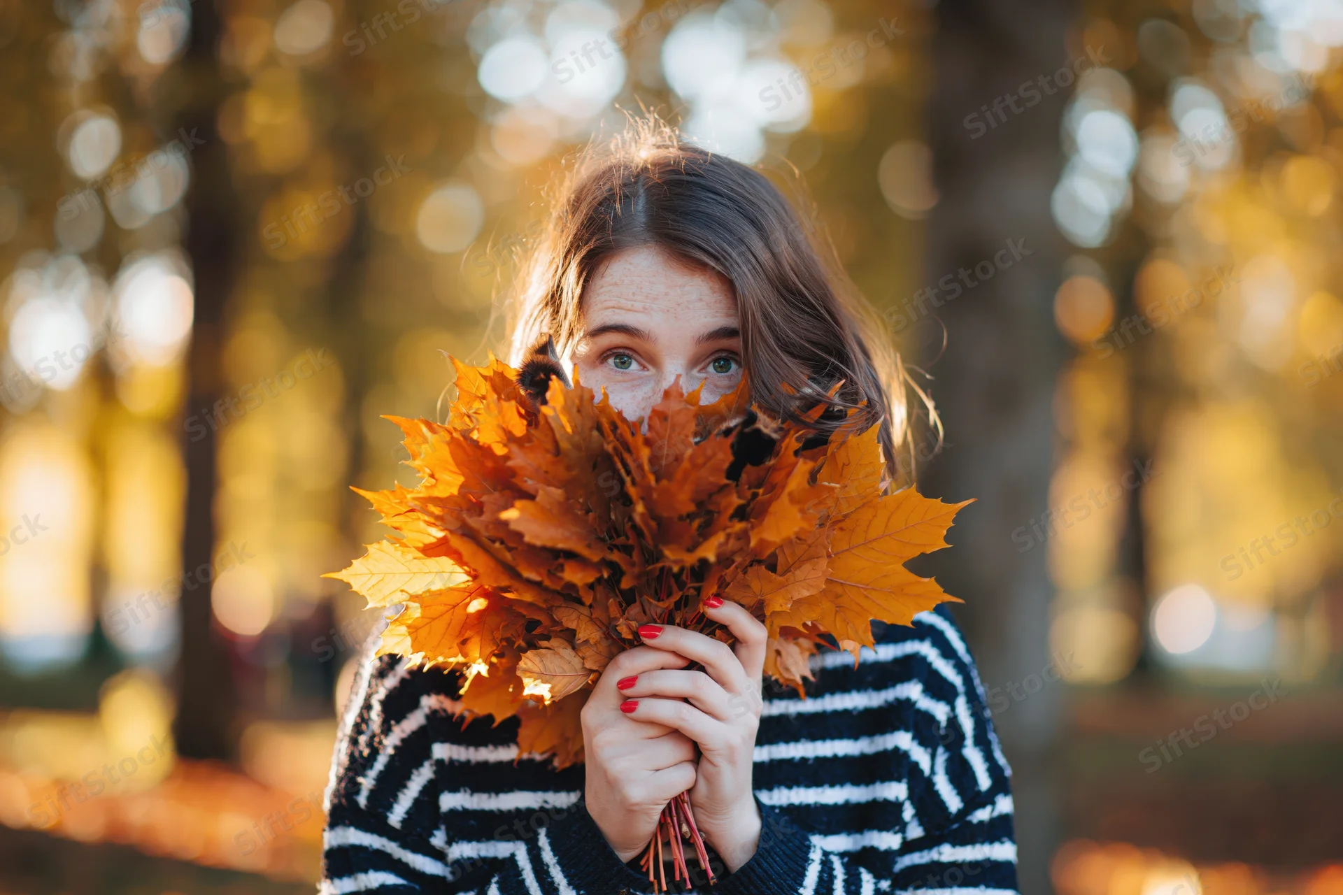 Young woman holding bouquet of orange maple leaves in front of her face in autumn park